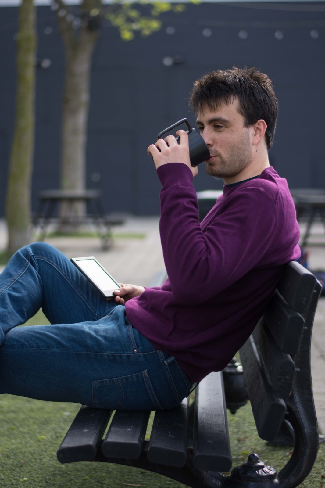Client session photo: man in a plum sweater relaxing on a park bench with a coffee and tablet