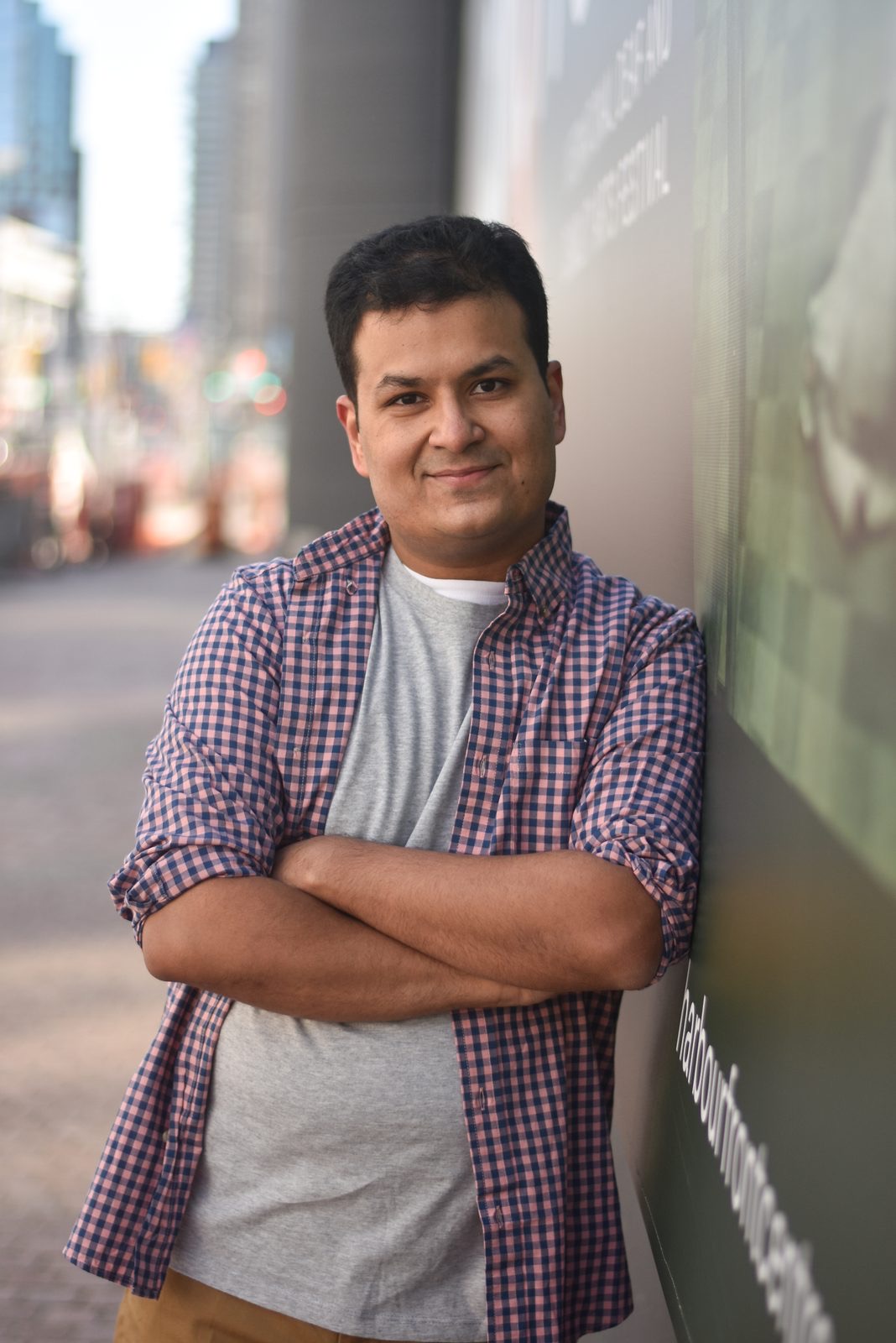 Client session photo: man in a plaid shirt with arms crossed leaning on a wall by a downtown street